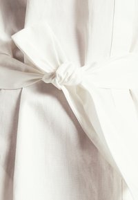 Tissu en lin blanc avec une ceinture nouée à la taille. La texture est douce et légère, avec un léger éclat, mettant en valeur un design élégant.