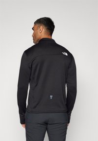 Veste noire à manches longues avec un motif texturé et un col montant. Dotée d'un logo blanc au dos et d'un discret accent bleu près de l'ourlet.