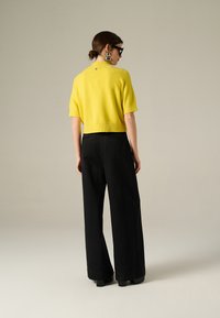 Maglione cropped giallo a maniche corte, abbinato a pantaloni neri a gamba larga. Il top presenta un piccolo dettaglio decorativo sul retro.