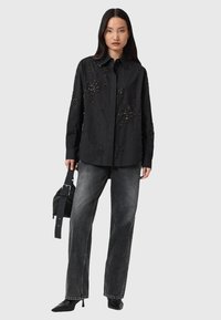 Camicia nera con chiusura a bottoni e decorazioni in perline, abbinata a jeans grigi a gamba larga e décolleté neri appuntiti. Include una borsa a tracolla nera.