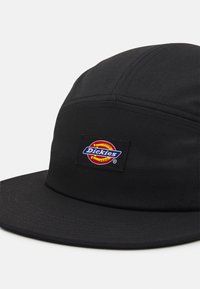 Černá kšiltovka s plochým kšiltem, vyrobená z texturované látky. Na ní je výrazně umístěna obdélníková nášivka s logem Dickies s barevným designem.