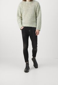 Persona vestida con un suéter de punto gris claro, jeans ajustados negros y botines negros, de pie con las manos en los bolsillos sobre un fondo blanco.