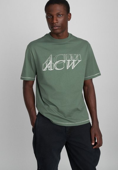 Grünes Baumwoll-T-Shirt mit auffälligem weißen Grafiketikett, kurzen Ärmeln, rundem Halsausschnitt und kontrastierendem Steppdetail.