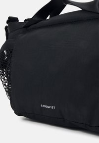 Borsa in tessuto nero con una tasca in rete texturizzata, cuciture rinforzate e un logo bianco. Presenta una tracolla regolabile e una superficie liscia.
