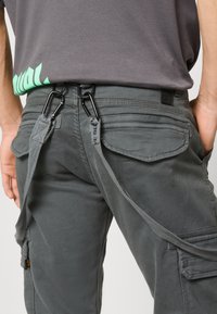 Pantalones cargo grises con un acabado texturizado, que cuentan con herrajes y correas negras sujetas a los lazos del cinturón para mayor utilidad.