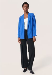 Erkblue blazers, mille sobiva lõikega, koos valge ribaga särgiga ja mustade laia jalgadega pükstega. Sile tekstuur ja selged jooned.