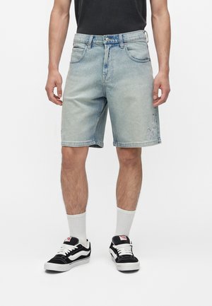 Mann trägt hellblaue Jeansshorts mit subtiler Sternstickerei, weiße Sneakersocken, schwarz-weiße Turnschuhe und ein schwarzes T-Shirt.