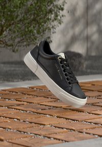 Μαύρο δερμάτινο αθλητικό παπούτσι με λευκή καουτσούκ σόλα, διαθέτοντας στρογγυλεμένη μύτη, επίπεδα κορδόνια και χρυσή σήμανση στο πλάι. Υφή στην επιφάνεια και κλασικός σχεδιασμός.