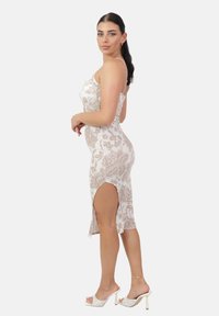 Donna con capelli scuri in un vestito midi floreale beige con spacco laterale, posando di lato indossando sandali con tacco bianchi e un sottile bracciale alla caviglia.