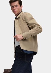 Beige lehká bunda s knoflíkovou přední stranou, jednou náprsní kapsou a dlouh�ými rukávy. Nosená přes šedou košili a tmavé džíny.