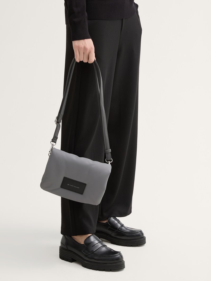 Personne portant un pantalon et des chaussures noirs tenant un petit sac à main gris avec une bandoulière noire ajustable et un écusson logo.
