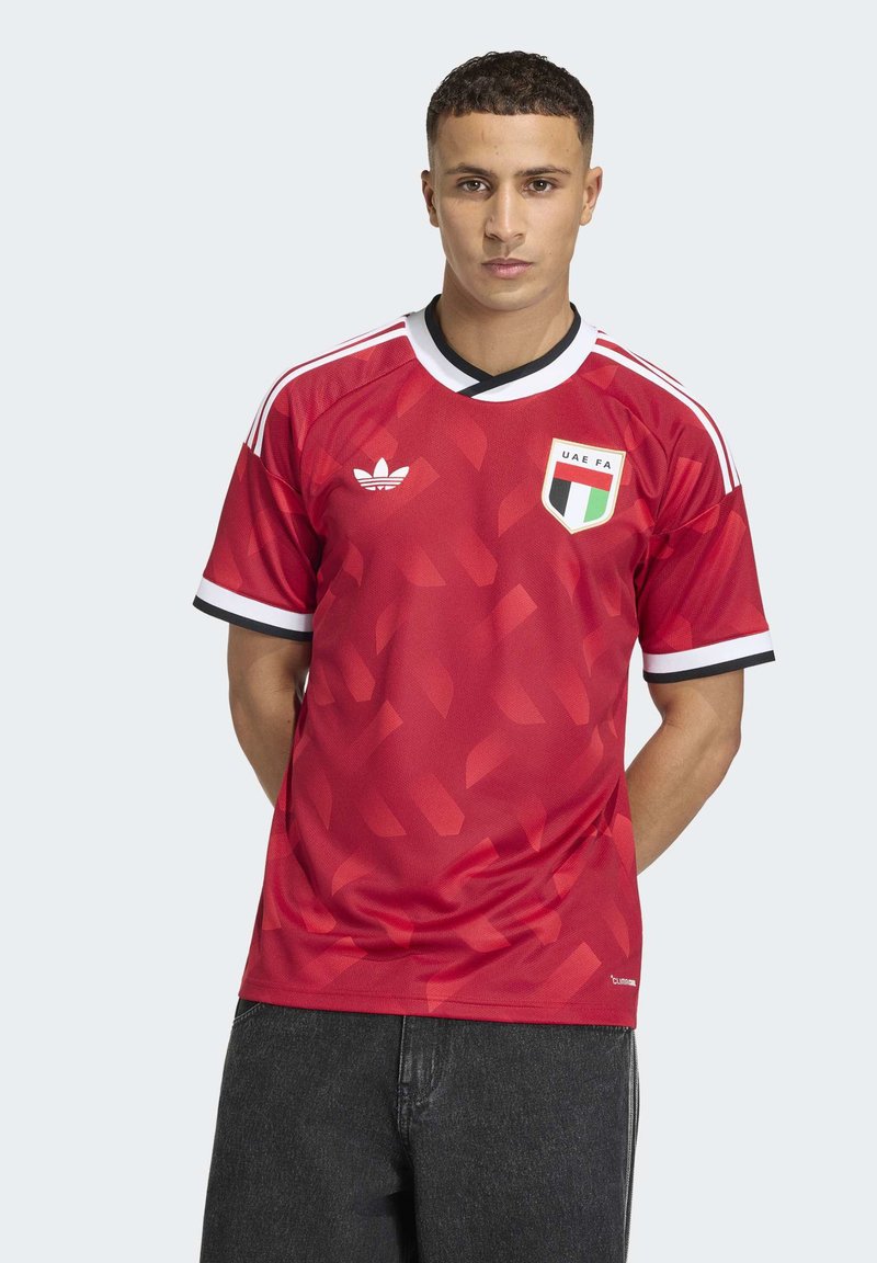 Jeune homme portant un maillot de football rouge de la Fédération de football des Émirats arabes unis avec des bordures blanches et noires, debout les mains derrière le dos sur un fond clair uni.