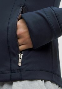 Jūras flotes krāsas aizdare ar rāvējslēdzēju un pieguļošu piedurkņu dizainu, ar sudraba rāvējslēdzēja detaļām un elegantu izskatu. Materiāls šķiet gluds un nedaudz teksturēts.