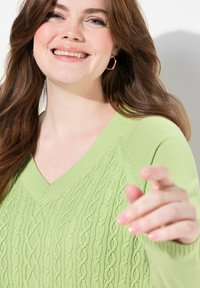 Vaaleanvihreä neulepusero, jossa on V-pääntie, kaapelikudontamallit ja ribbauskankaiset hihansuut. Malli ojentaa kätensä katsojaa kohti.