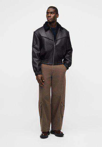 Άνδρας που φοράει μαύρο δερμάτινο μπουφάν με μαύρη επένδυση shearling στον γιακά, σκούφο πουλόβερ, καφέ παντελόνι cargo και σκούρα παπούτσια, στέκεται μπροστά σε μονόχρωμο γκρι φόντο.