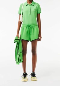 Polo vert, short assorti avec cordon de serrage, et un cardigan vert tenu à la main. Baskets noires et jaunes. Design décontracté avec une texture douce.