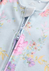 Floralni zip-up kombinezon od svijetloplavog materijala, koji sadrži cvjetne uzorke u ružičastoj, ljubičastoj i žutoj boji. Poliester u kombinaciji s mekanom teksturom.