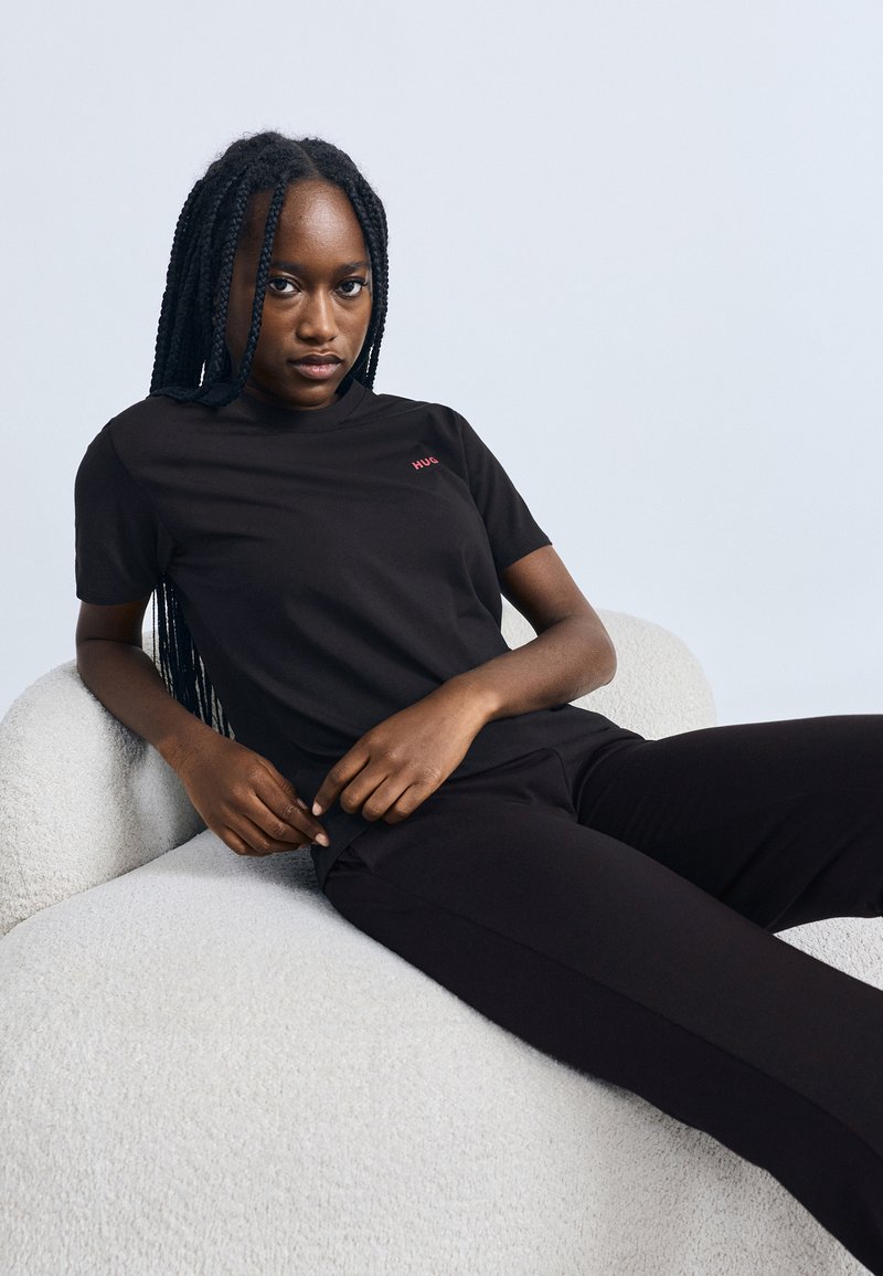 Fekete rövid ujjú póló piros "HUG" felirattal, fekete nadrággal párosítva. A modell egy világos textúrájú székben ül, fonott hajjal.