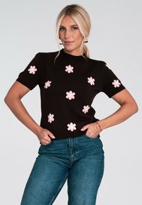 Blondynka w czarnym swetrze z krótkim rękawem w różowe kwiatowe wzory i niebieskich dżinsach, pozująca z rękami w kieszeniach na białym tle.