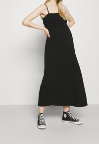 Henkilö, jolla on yllään hihaton musta maksimekko ja mustat korkeavartiset Converse-tennarit, seisoo kädet lantiolla yksinkertaista taustaa vasten.