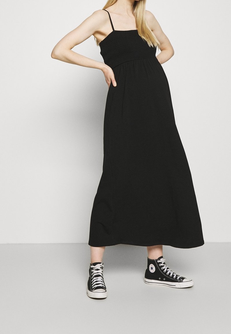 Henkilö, jolla on yllään hihaton musta maksimekko ja mustat korkeavartiset Converse-tennarit, seisoo kädet lantiolla yksinkertaista taustaa vasten.