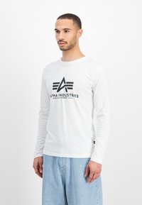 Bijela majica dugih rukava s crnim logotipom koji glasi "Alpha Industries" s geometrijskim naglascima na prednjem dijelu.