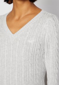 Pull en tricot gris clair avec un col en V, un design côtelé avec des motifs en torsades et des manches longues. Texture douce avec un léger éclat.