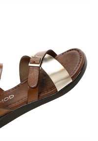 Sandal in pelle marrone con un accento metallico dorato sulla parte anteriore, fibbia regolabile, soletta ammortizzata e suola nera con texture.