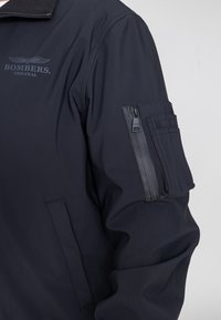 Marineblaue Bomberjacke mit einer Reißverschlusstasche am linken Ärmel, ausgestattet mit einer glatten Textur und gesticktem Logo auf der Brust.