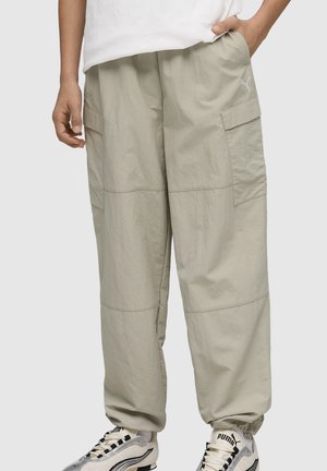 Persoon die lichtbeige cargobroek en witte Puma-sneakers draagt, staande met één hand in de zak tegen een effen achtergrond.