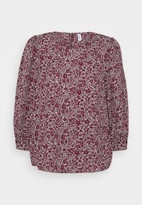 Blouse à manches longues avec motif floral violet sur fond blanc, encolure ronde et manches légèrement bouffantes.