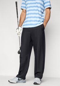 Μαύρο παντελόνι γκολφ με λεία υφή, σχεδιασμένο με άνετη εφαρμογή. Συνδυασμένο με μπλε ριγέ πόλο πουκάμισο και γκρι αθλητικά παπούτσια.