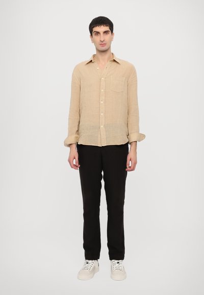Camicetta button-up in lino beige con taschino sul petto, maniche arrotolate, abbinata a pantaloni neri e sneakers bianche. Design minimalista.