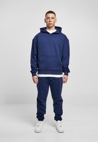 Mørkeblå hettegenser og matching joggebukser laget av myk stoff; har frontlomme, oversized passform og hvite sko.
