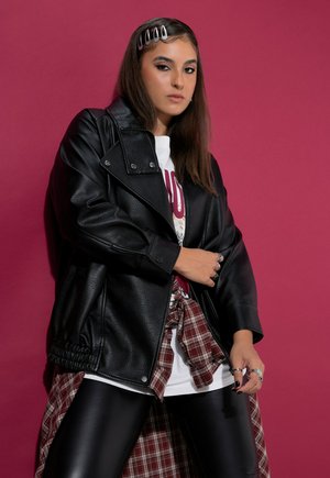Una mujer posa frente a un fondo magenta con una chaqueta de cuero negra, una camiseta blanca estampada, una camisa de cuadros atada a la cintura y unos leggings negros.