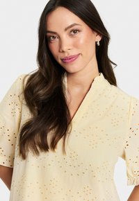 Gul broderad blus med korta puffärmar, med utskurna blommönster och v-ringning. Slät, lätt tyg.