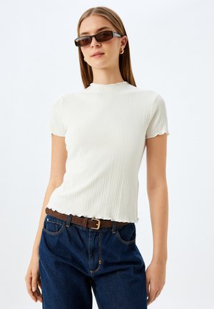 Top blanco de manga corta con relieve y escote festoneado, combinado con jeans de mezclilla azul oscuro y un cinturón marrón con hebilla dorada.