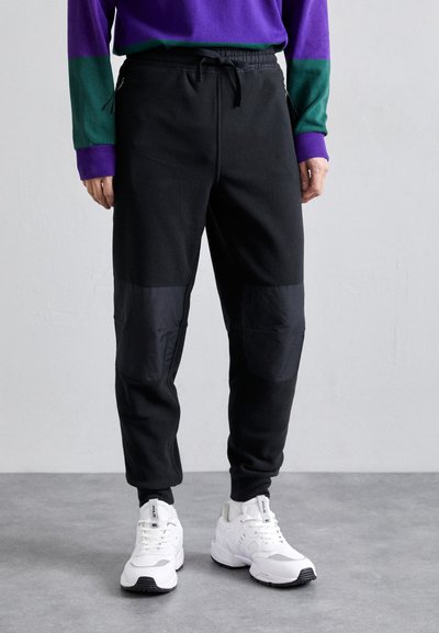 Černé joggers s tapered střihem, s texturovanými kolenními panely a elastickým pasem. Spárováno s bílými sportovními botami.