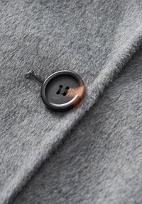 Grauer Stofftextur mit einer Nahaufnahme eines dunklen runden Knopfes mit zwei Löchern und glänzender Oberfläche, der mit einer Faden-Schlaufe befestigt ist.