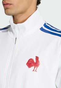 Biała kurtka na zamek z niebieskimi paskami na ramionach i wyraźnym czerwonym haftowanym logo koguta na piersi.