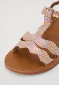 Sandali rosa chiaro con cinturini ondulati e iridescenti, caratterizzati da un motivo testurizzato e una suola piatta marrone. Dettagli cuciti lungo i bordi.