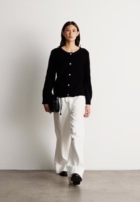 Juoda megzta kardiganas su apvaliu kaklu ir mygtukų užsegimu, derinamas su baltais plačiais kelnėmis ir maža juoda rankine. Modelis vilki plokščius batus.
