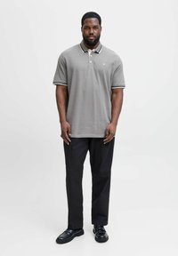 Man staat op een witte achtergrond en draagt een grijze polo, zwarte broek en zwarte schoenen. Hij kijkt vooruit met ontspannen handen.