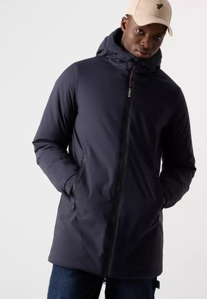 Hombre con chaqueta con capucha oscura, manos en los bolsillos y una gorra de béisbol beige claro sobre un fondo liso.