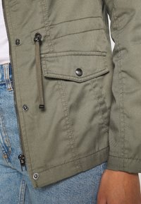 Näruppfattning av en olivgrön jackficka med knappklapp, dragkedja och justerbar dragsko, ovanpå blå jeans och en hand som vilar bredvid.