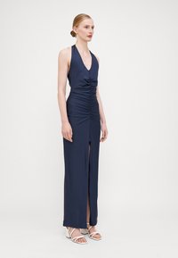Mørkeblå halter maxi kjole laget av glatt stoff, med rynking foran og høy splitt til låret. Brukt med hvite sandaletter.