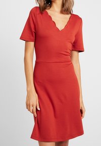 Žena v upnutých červených šatech s krátkými rukávy a výstřihem do V s vlnitým okrajem, stojící proti jednoduchému světlému pozadí.