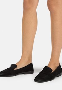 Mocassini neri in suede con una parte anteriore strutturata, indossati ai piedi. Presentano una punta arrotondata e un tacco piatto con un design semplice.