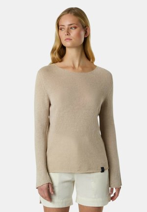 Donna con lunghi capelli biondi che indossa un maglione beige a maniche lunghe e pantaloncini color crema, in piedi davanti a uno sfondo semplice.