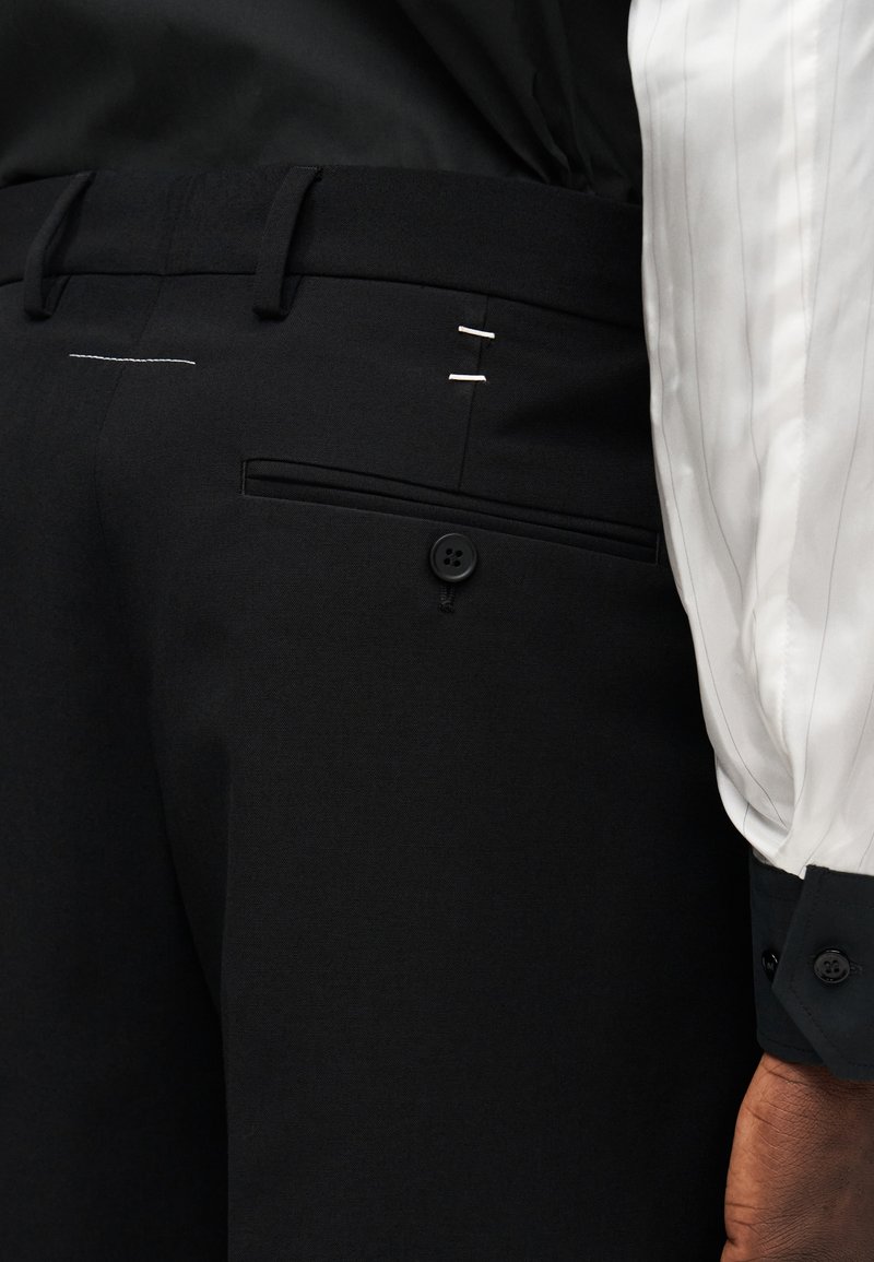 Pantalon noir ajusté avec une texture lisse, doté d'une fermeture à bouton, de deux accents cousus et d'une poche arrière.